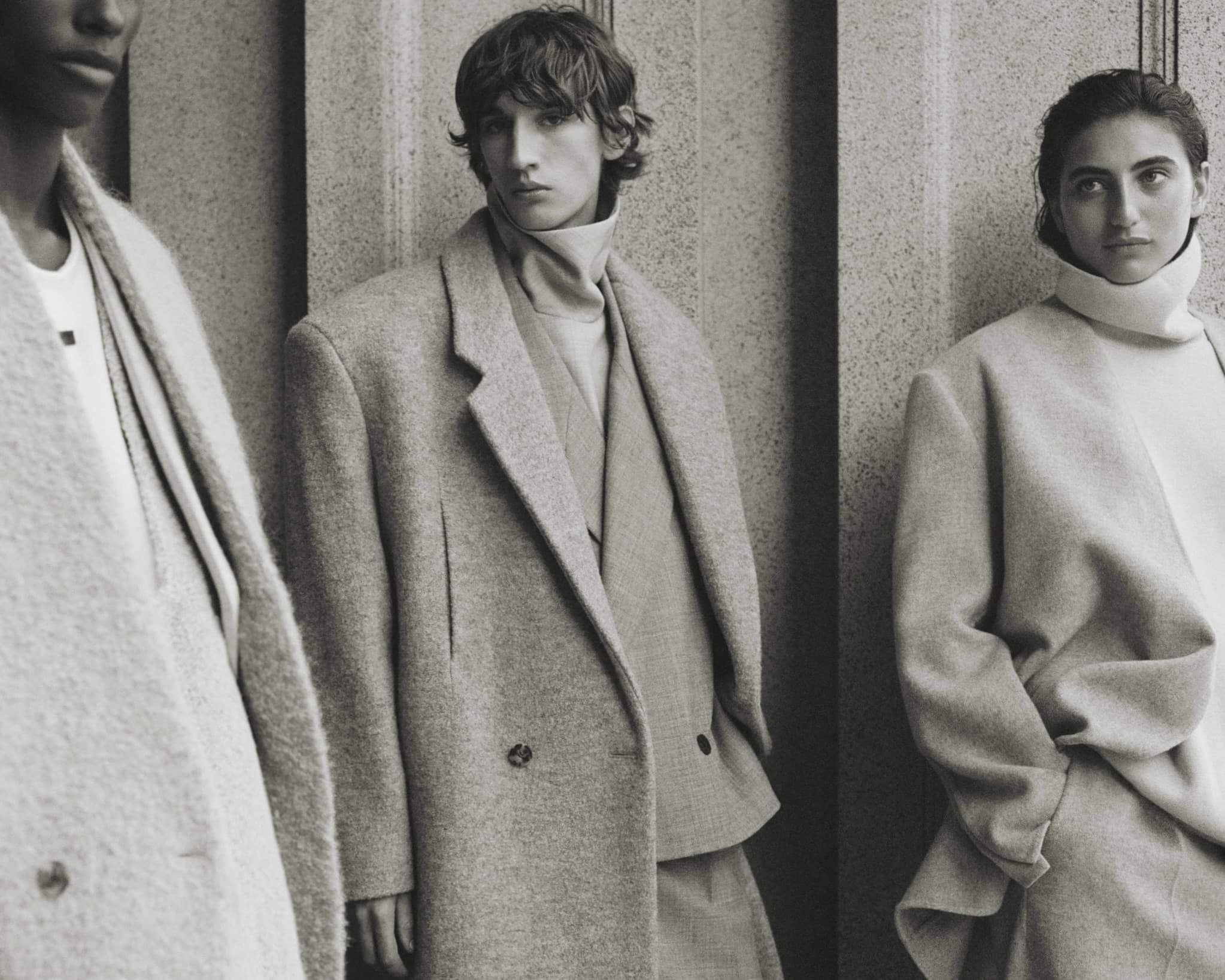Three models wearing structured neutral coats and turtlenecks posed against a textured stone architectural wall.