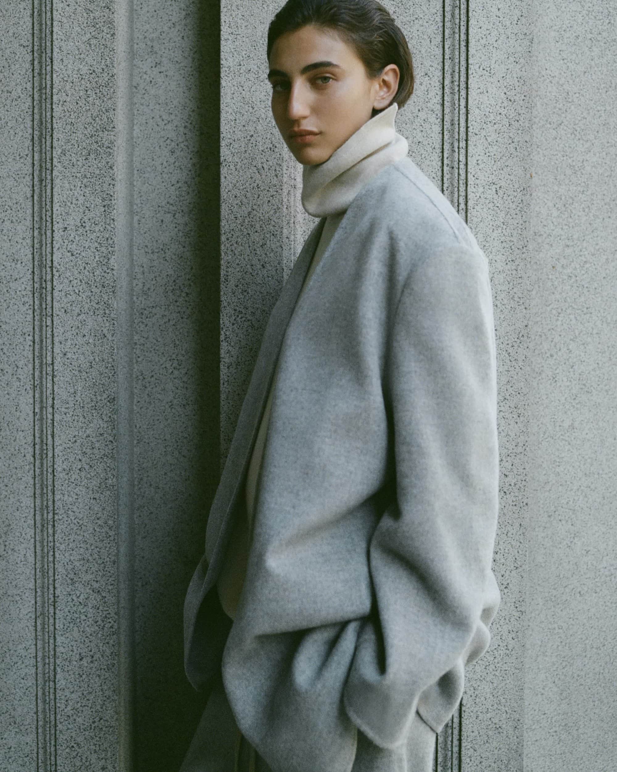 Model wearing a grey wool coat and turtleneck standing against a textured stone architectural pillar.