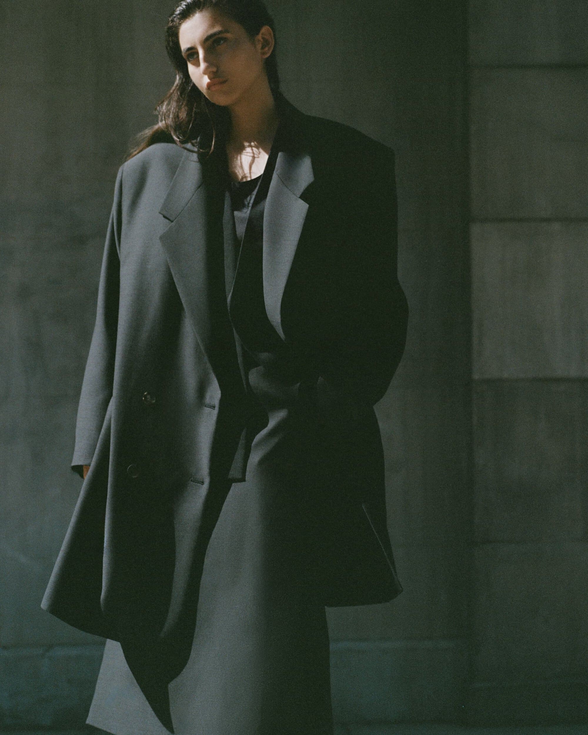 Model standing in a long dark structured coat and matching skirt against a concrete wall in natural daylight.
