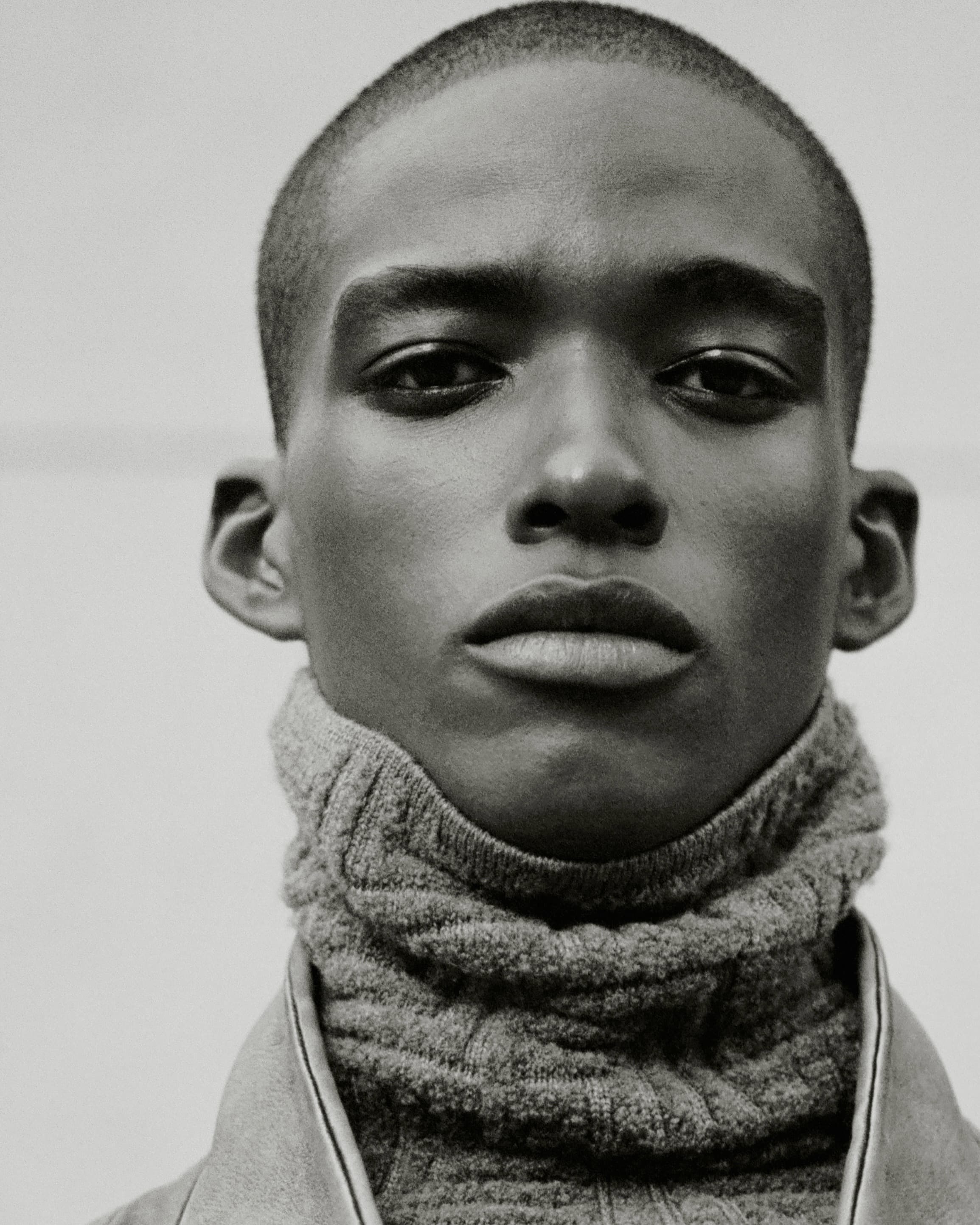 Close-up portrait of a man wearing a high-neck textured grey sweater against a plain background.