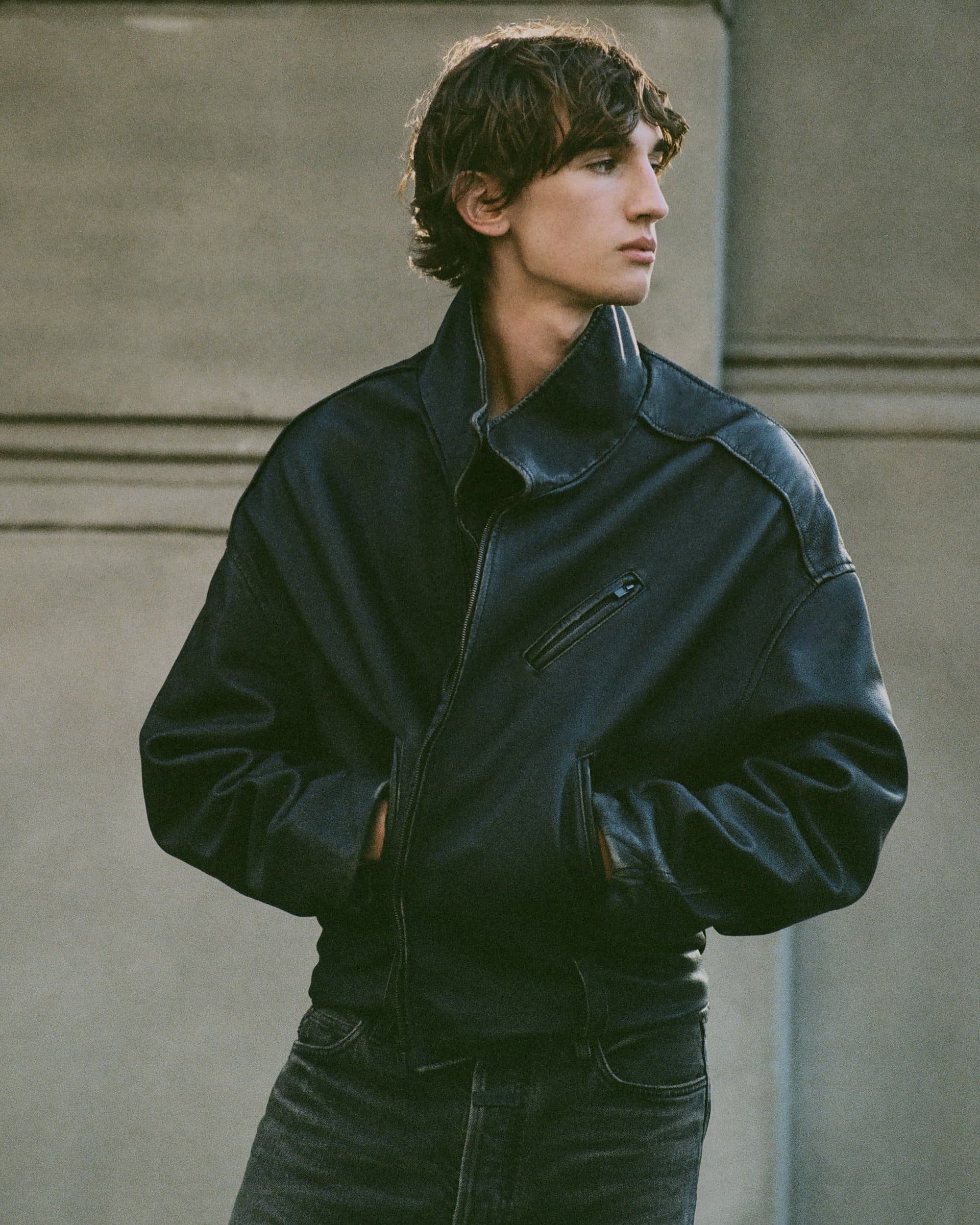 Young male model wearing a dark leather jacket with a high collar and dark denim jeans against an exterior wall