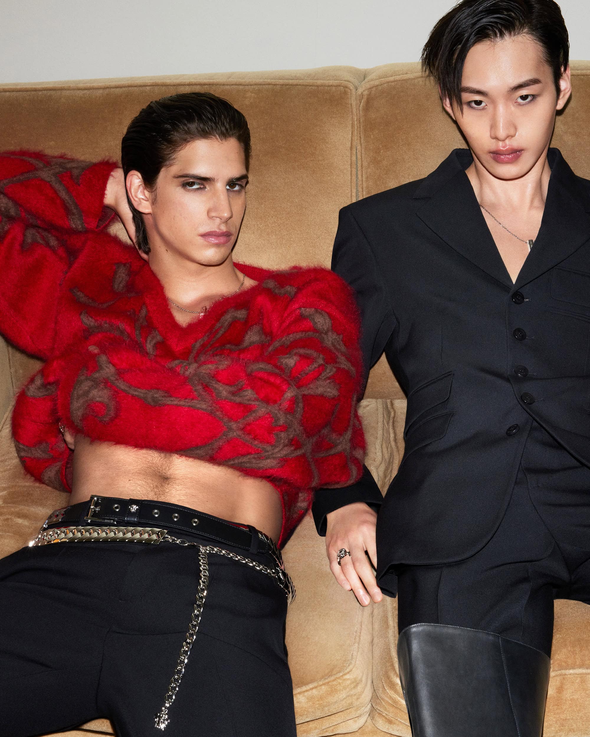 Two male models posing on a tan couch, one wearing a red patterned sweater and black trousers, the other in a black suit.