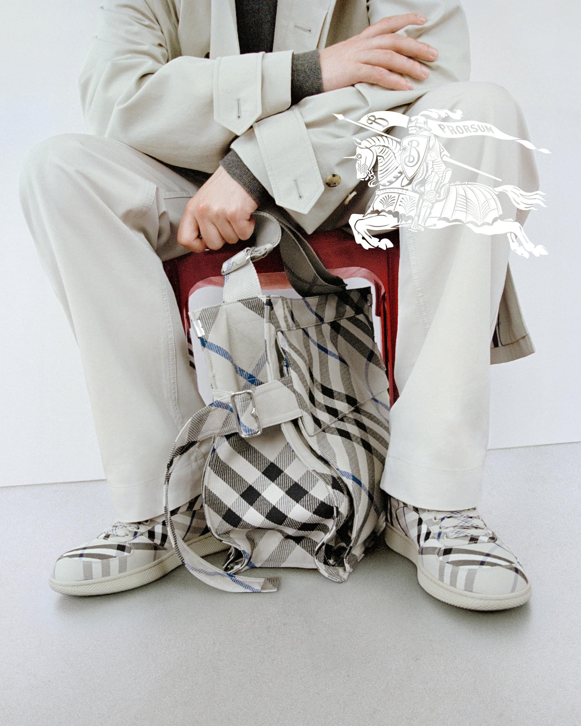 Seated model wearing beige trench coat and plaid sneakers holding a matching plaid tote bag in a studio setting