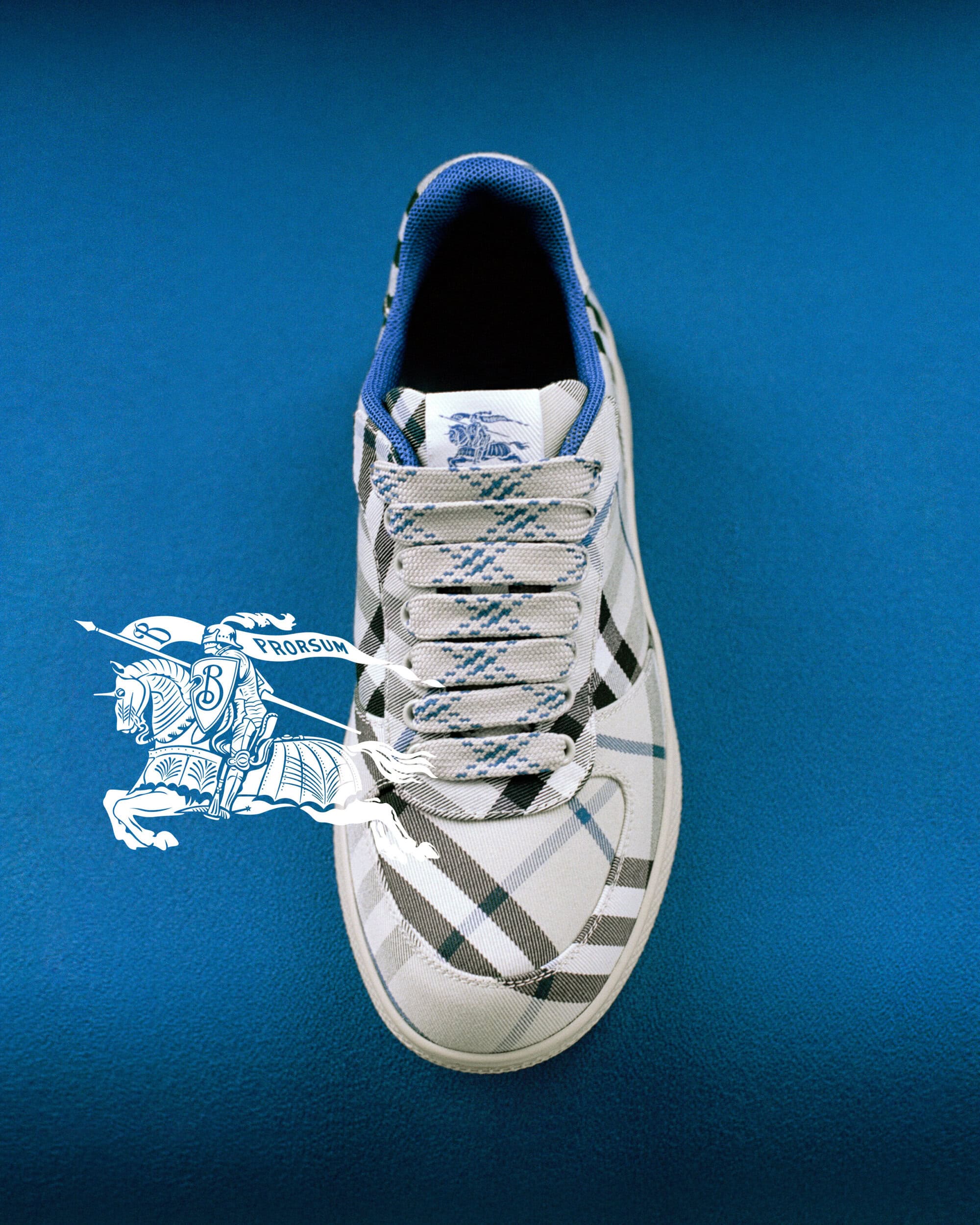 Top-down view of a Burberry plaid sneaker on a solid blue surface with a white knight logo overlay.