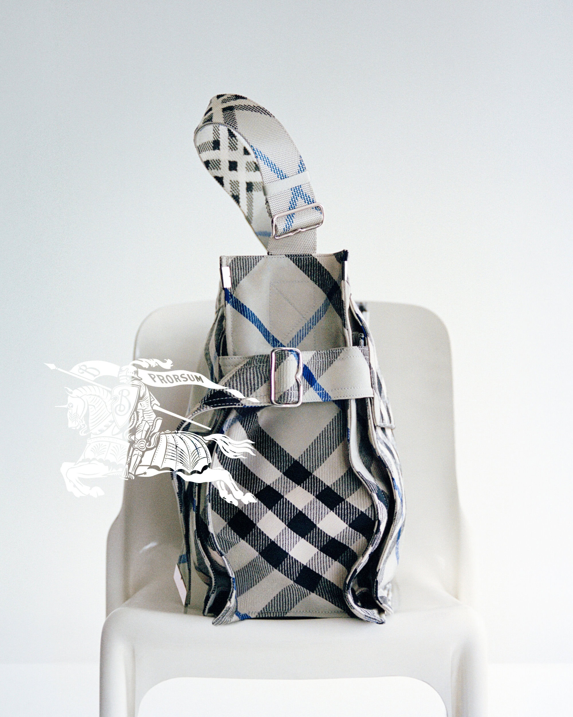Burberry plaid handbag resting on a white chair against a plain white background with a faint equestrian knight logo.