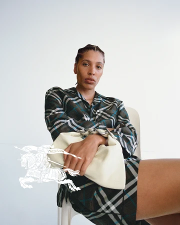 Model wearing a plaid shirt and holding a cream handbag seated against a plain white background in a Burberry campaign