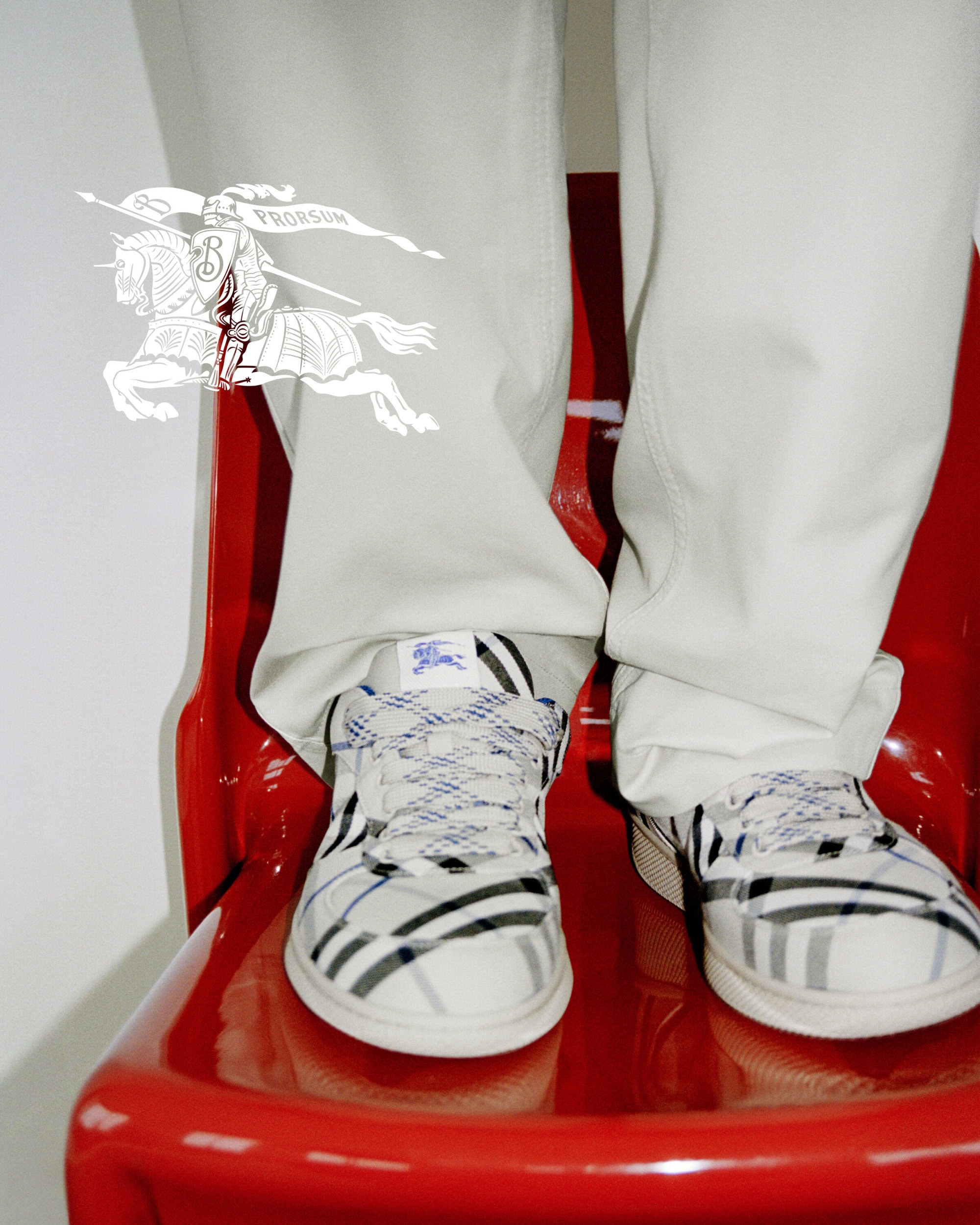 Close-up of person wearing Burberry plaid sneakers and light trousers, standing against a white backdrop.