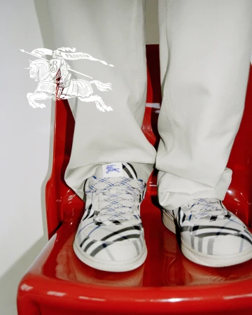 Close-up of person wearing Burberry plaid sneakers and light trousers, standing against a white backdrop.