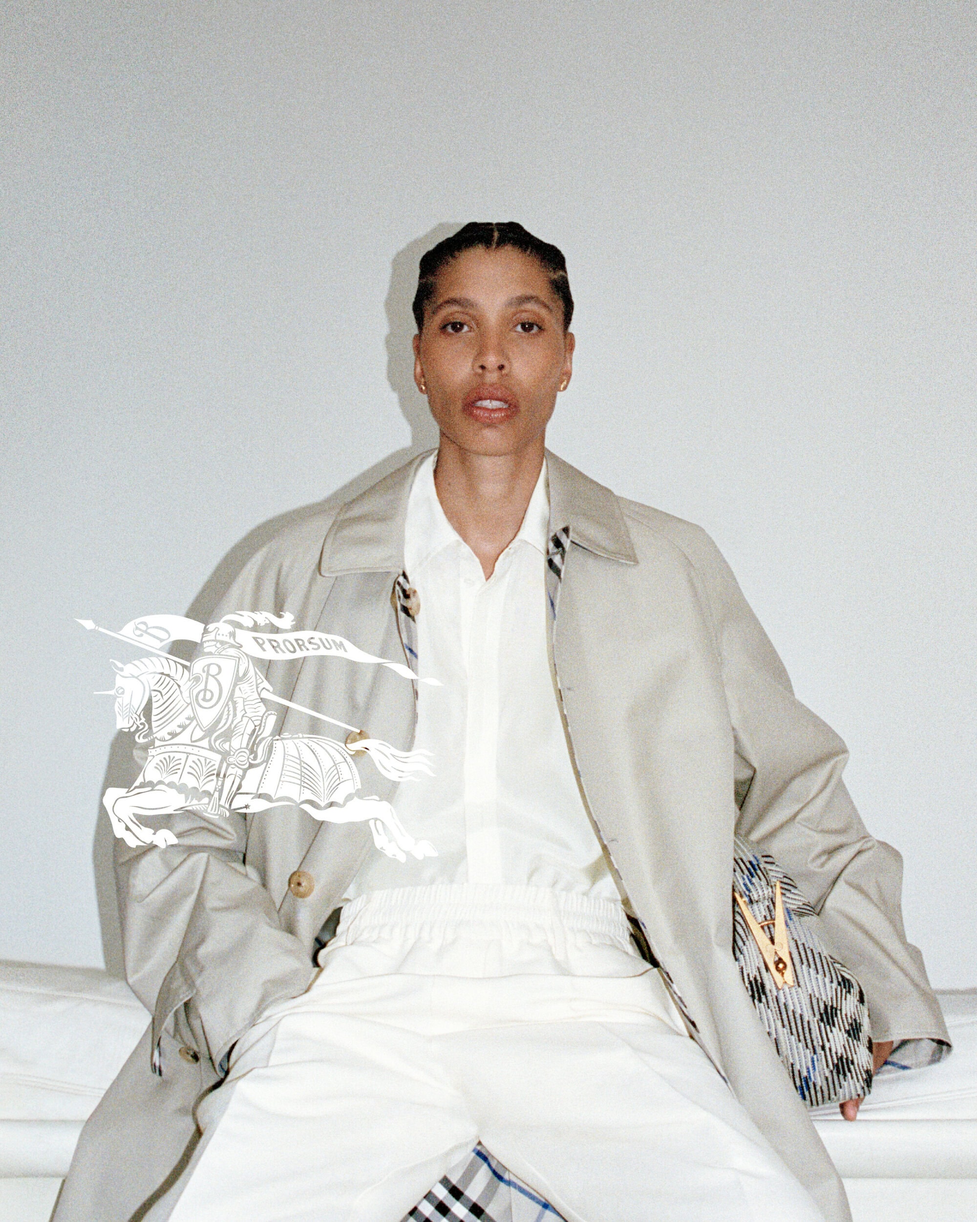 Model wearing light trench coat and white trousers holding patterned bag in front of white wall