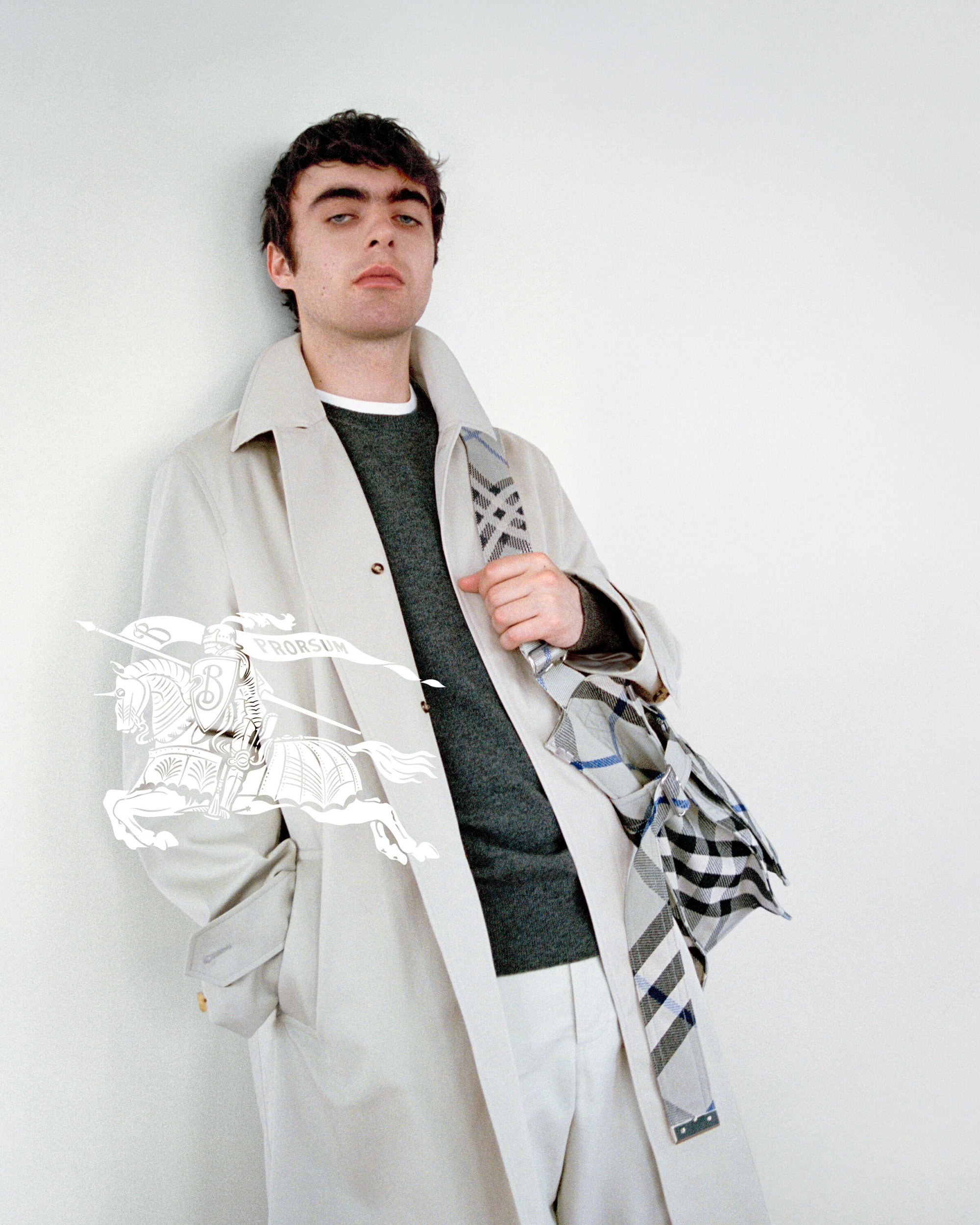 Man leaning against a white wall wearing a beige trench coat, dark green sweater, and holding a plaid umbrella for Burberry.