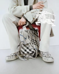 Seated model wearing beige trench coat and plaid sneakers holding a matching plaid tote bag in a studio setting