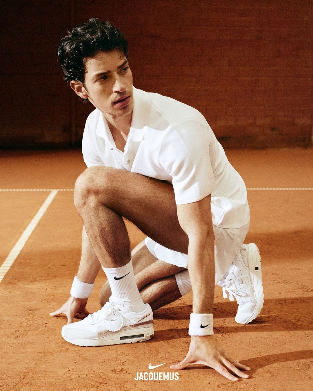 Male model in white polo shirt and shorts crouching on a clay tennis court wearing branded sneakers and wristbands