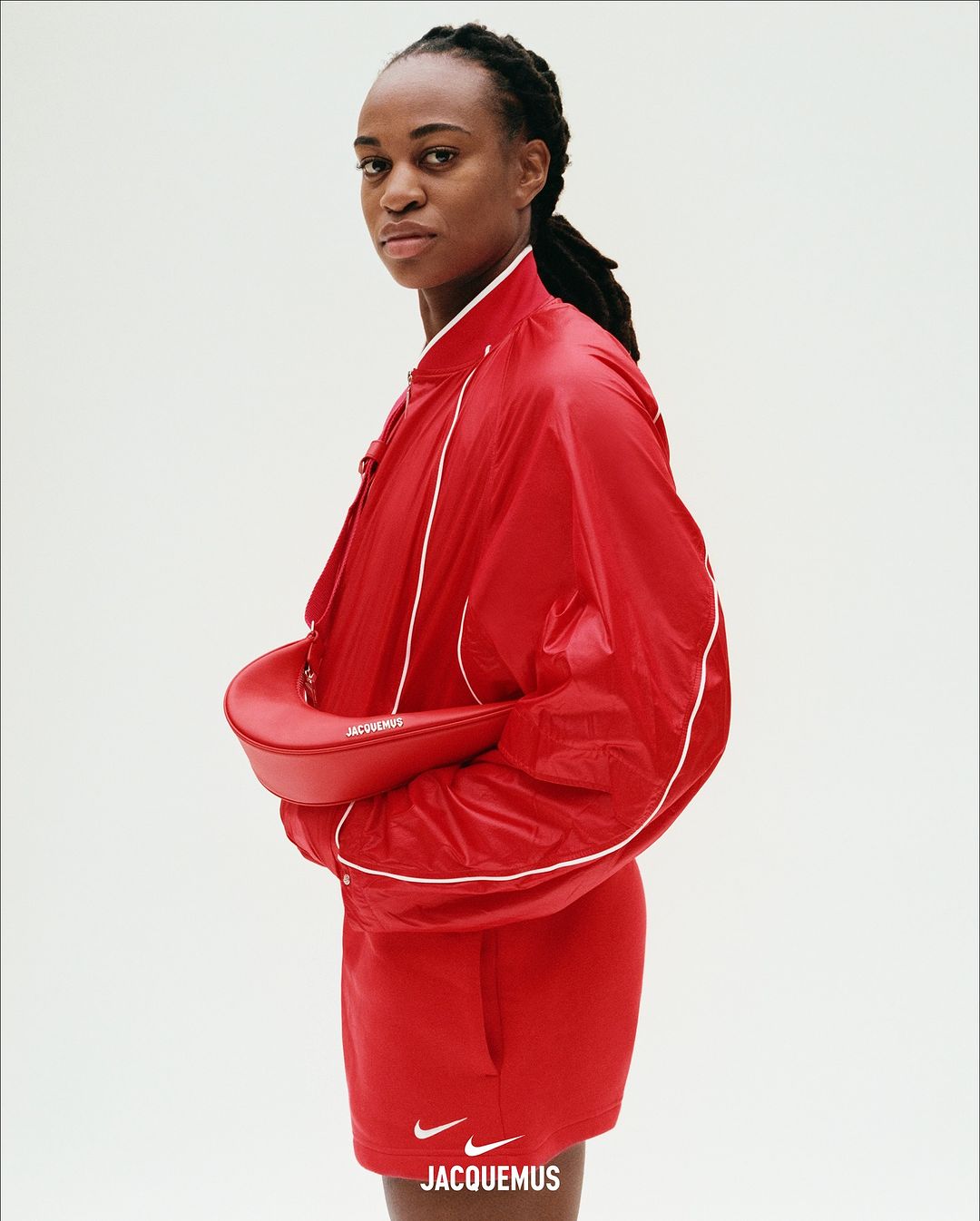 Woman wearing a bright red zip jacket and holding a matching red crescent shoulder bag against a plain white studio background.