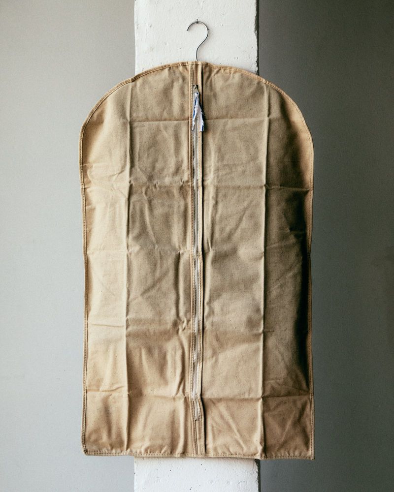 Tan garment bag hanging from a metal hanger against a gray wall with a white structural column in a studio setting.