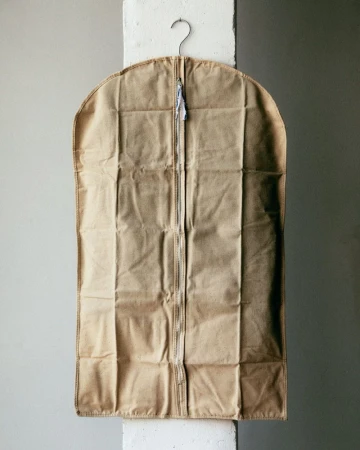 Tan garment bag hanging from a metal hanger against a gray wall with a white structural column in a studio setting.
