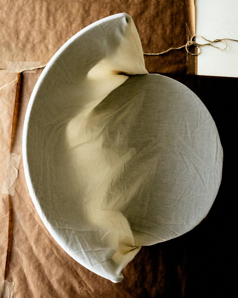 Top-down view of a round baking mold covered with cloth, on a brown paper surface with natural light creating shadows and highlights.