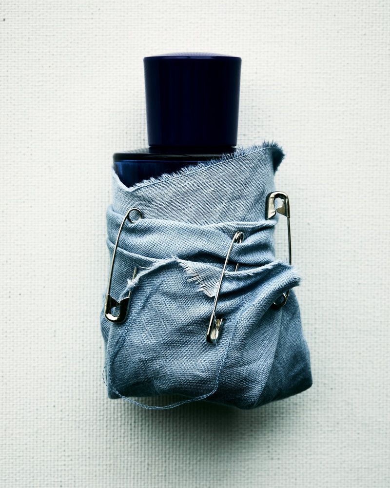 Elevated shot of a dark blue bottle wrapped in light blue cloth secured by safety pins against a textured off-white backdrop.