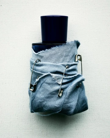 Elevated shot of a dark blue bottle wrapped in light blue cloth secured by safety pins against a textured off-white backdrop.