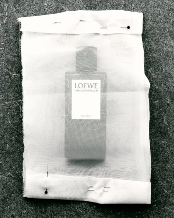 Still-life product shot of a Loewe perfume bottle encased in translucent fabric, pinned to gray textured surface, capturing minimalist aesthetic.
