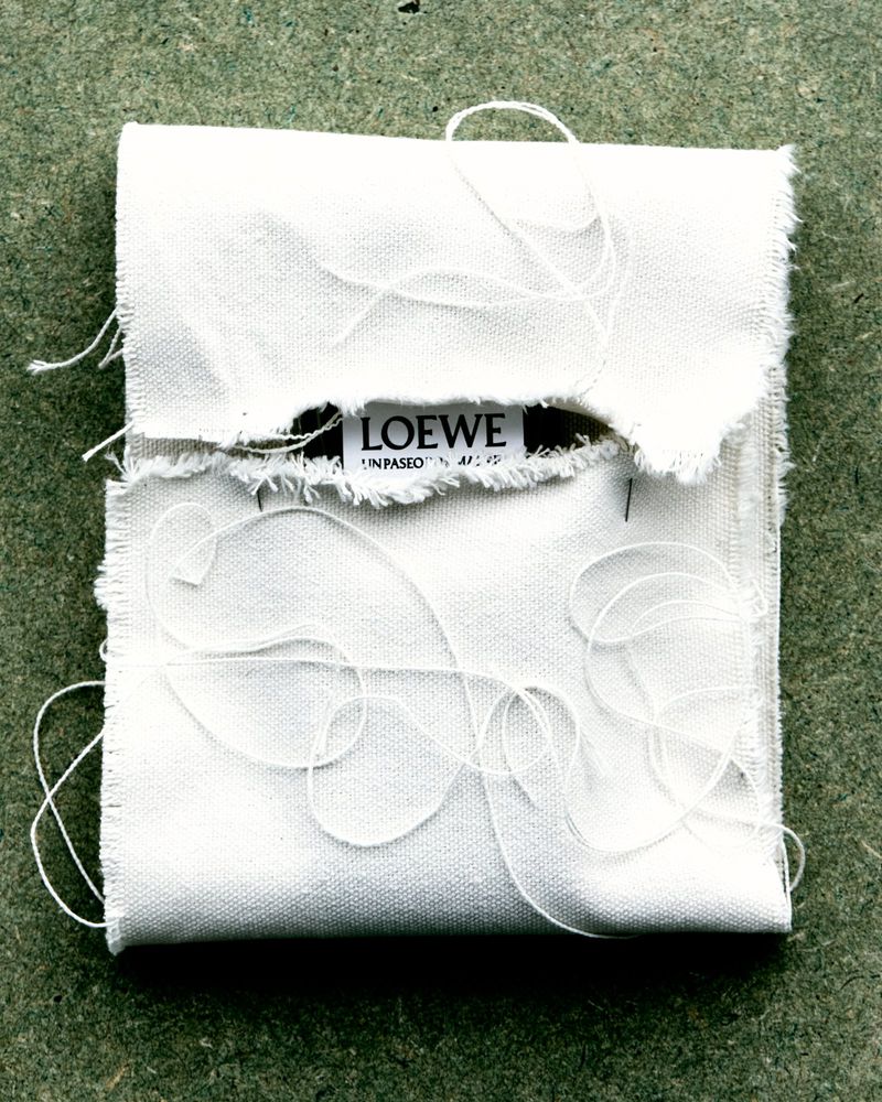 Close-up of a frayed-edge white fabric pouch with loosely-placed threads, bearing the 'LOEWE' logo against a neutral background.