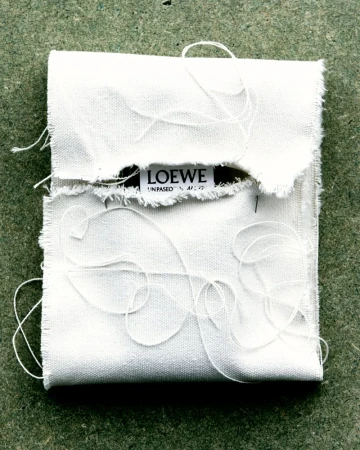 Close-up of a frayed-edge white fabric pouch with loosely-placed threads, bearing the 'LOEWE' logo against a neutral background.