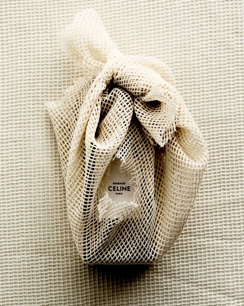 Celine branded item wrapped in a white mesh bag on a textured white surface. Soft, diffused lighting, neutral color palette.