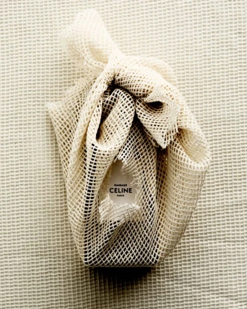 Celine branded item wrapped in a white mesh bag on a textured white surface. Soft, diffused lighting, neutral color palette.