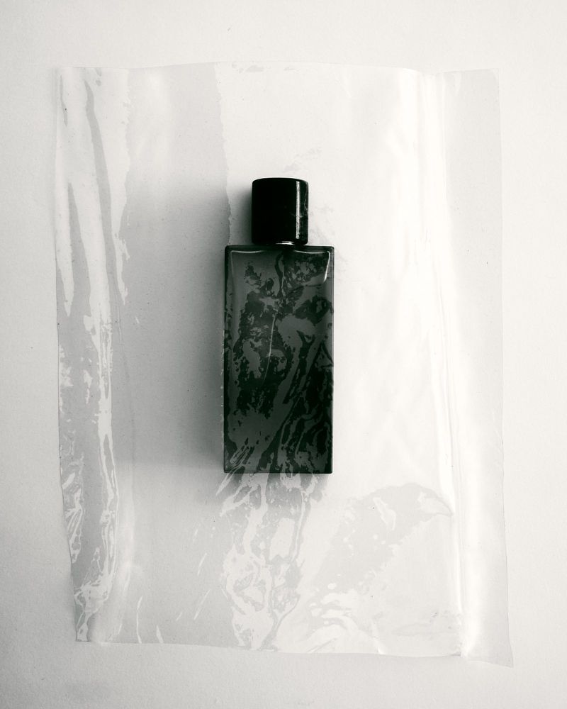 Close-up studio shot of a smoky green perfume bottle inside a clear plastic bag against a white background. Minimalist aesthetic.