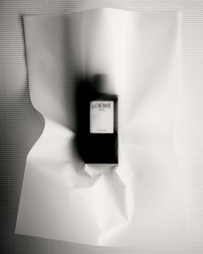 Studio shot of a bottle of Loewe Solo perfume wrapped in white paper against a horizontally ribbed, light grey wall.