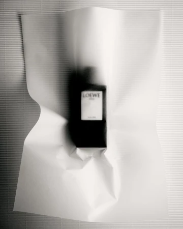 Studio shot of a bottle of Loewe Solo perfume wrapped in white paper against a horizontally ribbed, light grey wall.