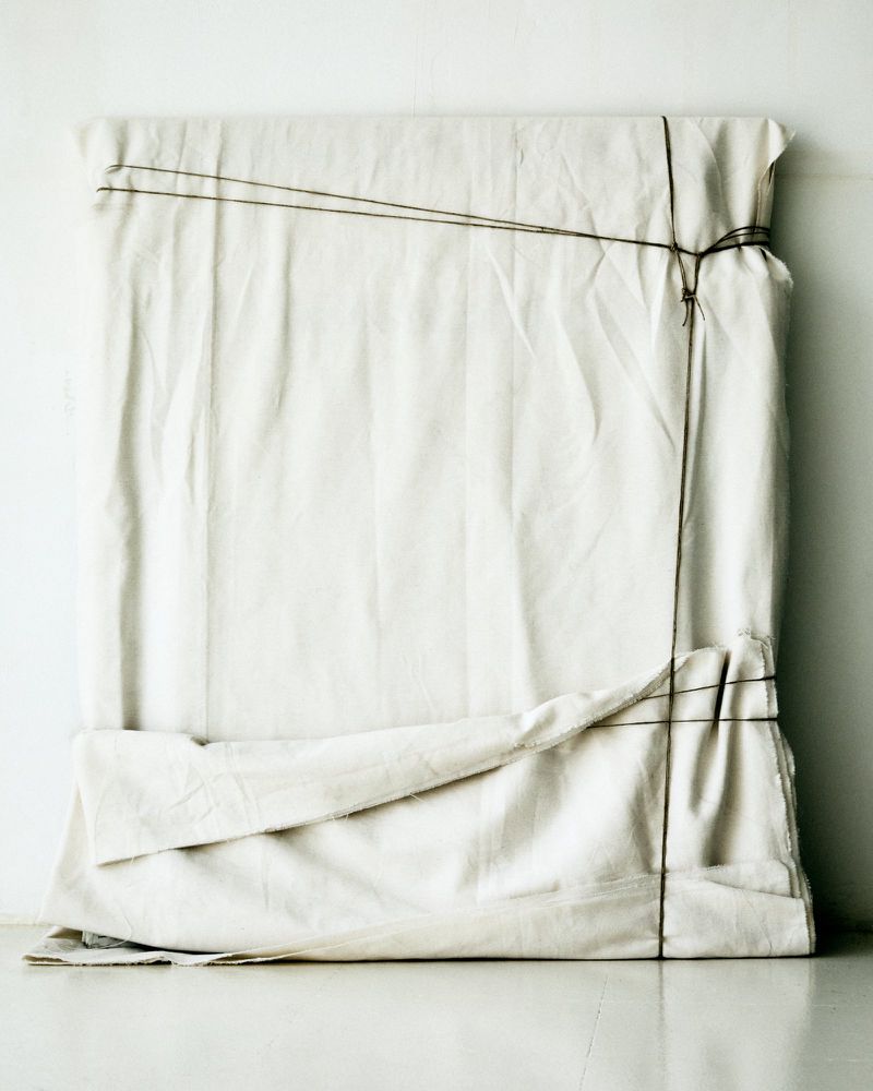 White cloth-covered object with dark string tied around it sitting on a white surface against a white wall. Neutral and minimal.