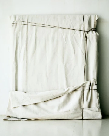 White cloth-covered object with dark string tied around it sitting on a white surface against a white wall. Neutral and minimal.