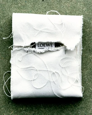 Folded white canvas fabric with frayed edges and loose threads, showing a small Loewe brand label against a textured green background