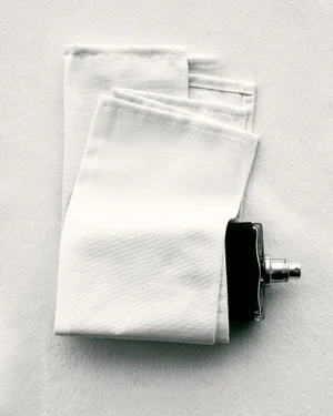 Folded white fabric cloth resting next to a dark perfume bottle with a silver spray nozzle on a textured white background.