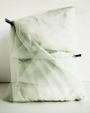 White pillows wrapped in translucent sage green tulle fabric fastened with black binder clips against a plain background.