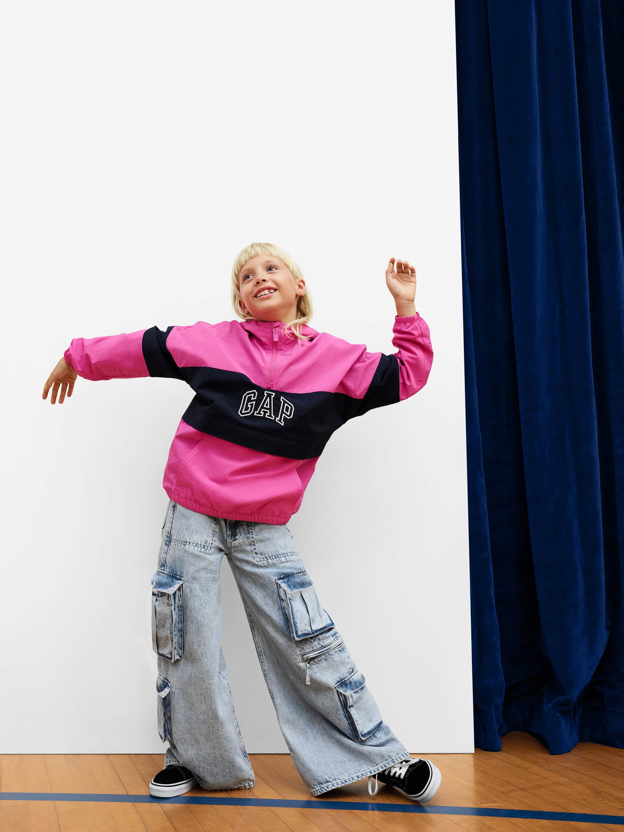 Child in pink and black windbreaker and cargo pants dancing against a white wall with blue curtains.