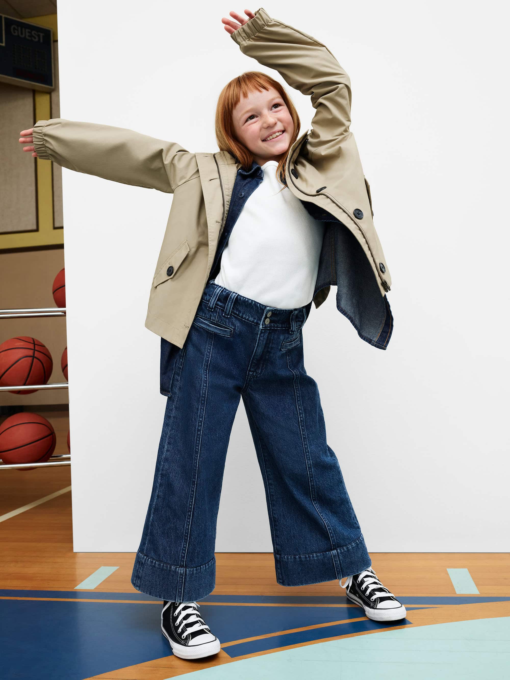 Young red-haired girl wearing beige jacket and blue jeans dancing against white studio background
