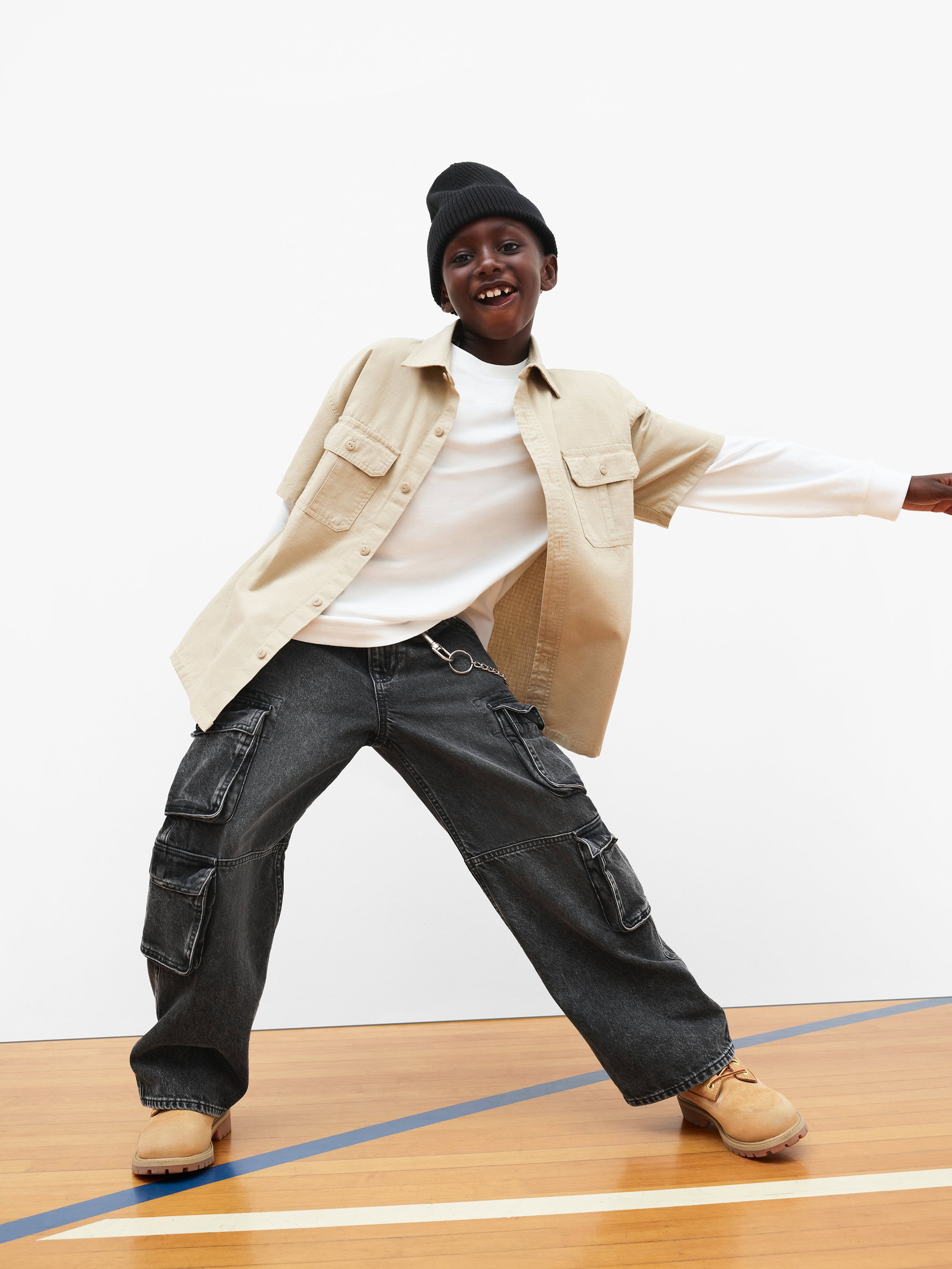 Child in black beanie, beige overshirt and dark cargo pants posing dynamically on a wooden floor.