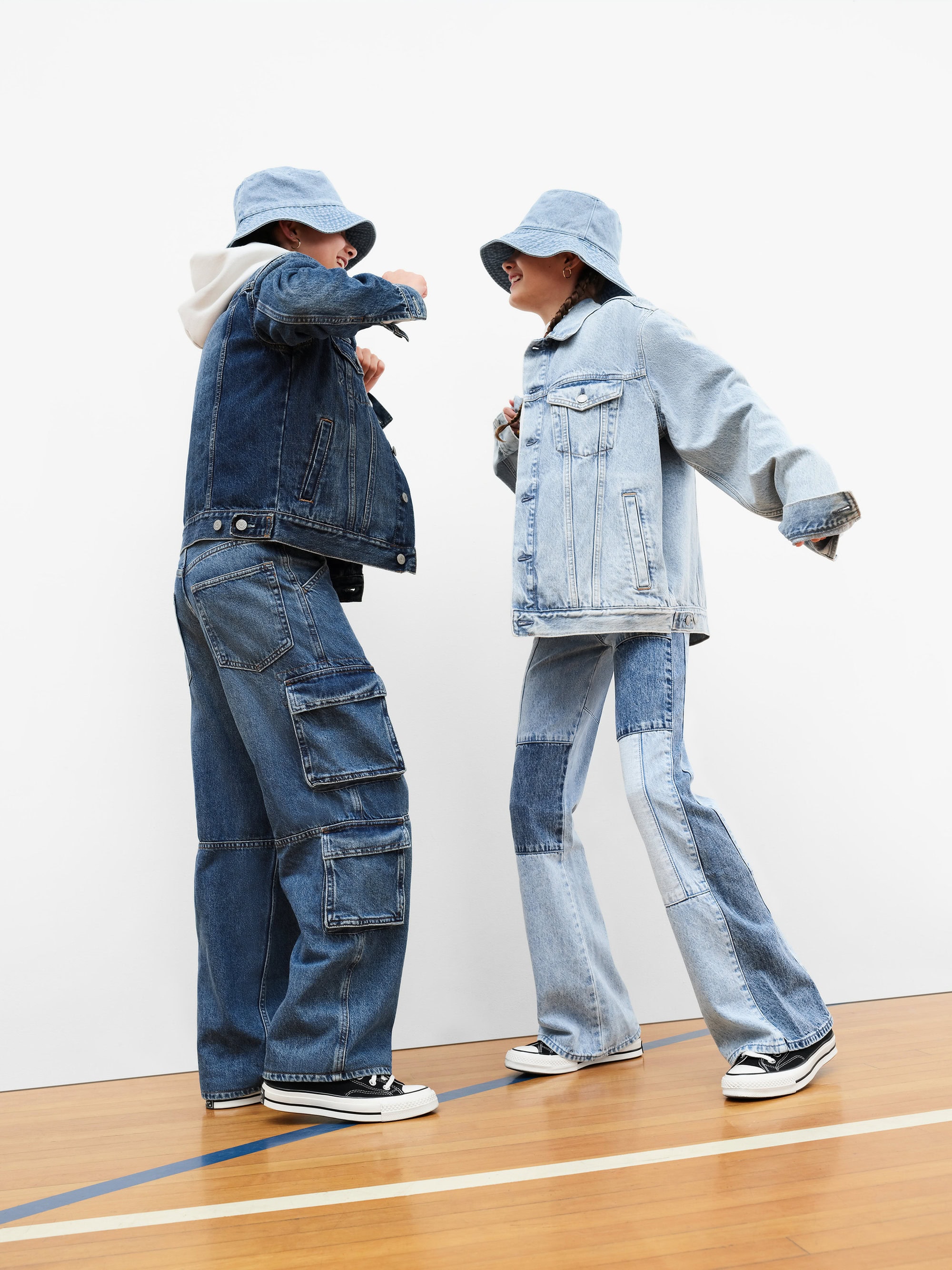 Two people wearing matching denim jackets and bucket hats dancing on a polished wooden floor against a white background.