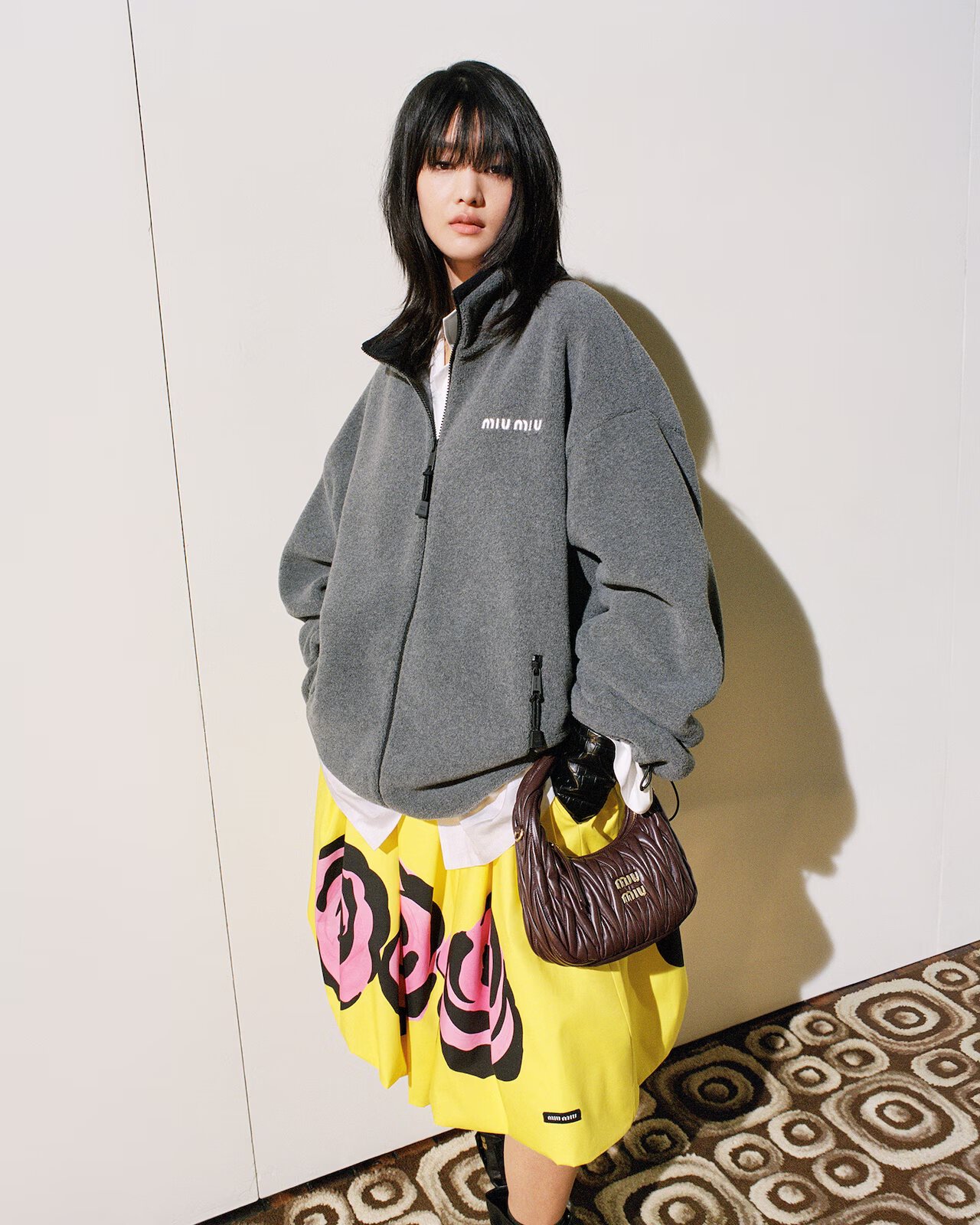 Model wearing a grey fleece jacket, yellow patterned skirt, and holding a brown shoulder bag against a plain wall.