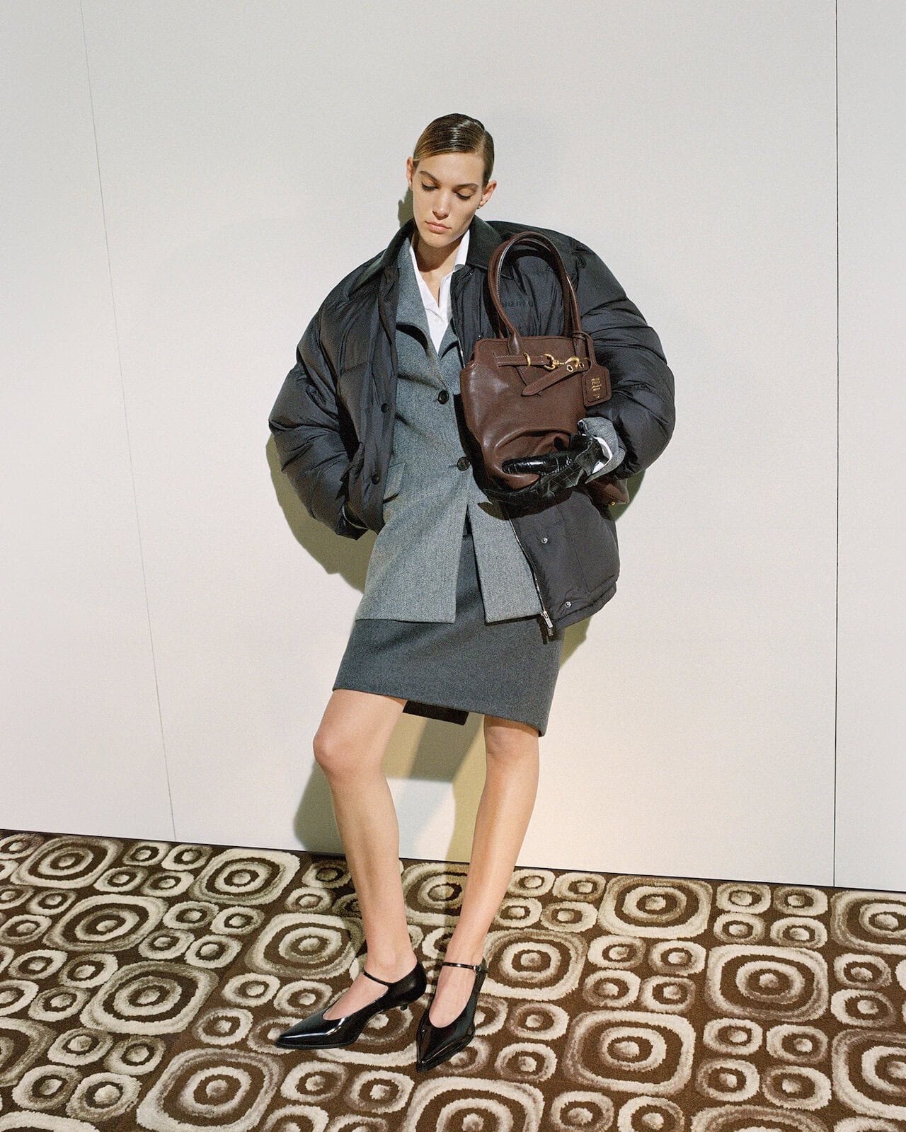 Fashion model poses in a gray skirt suit and black puffer jacket, accessorized with a brown purse and black Mary Jane shoes on a patterned floor.