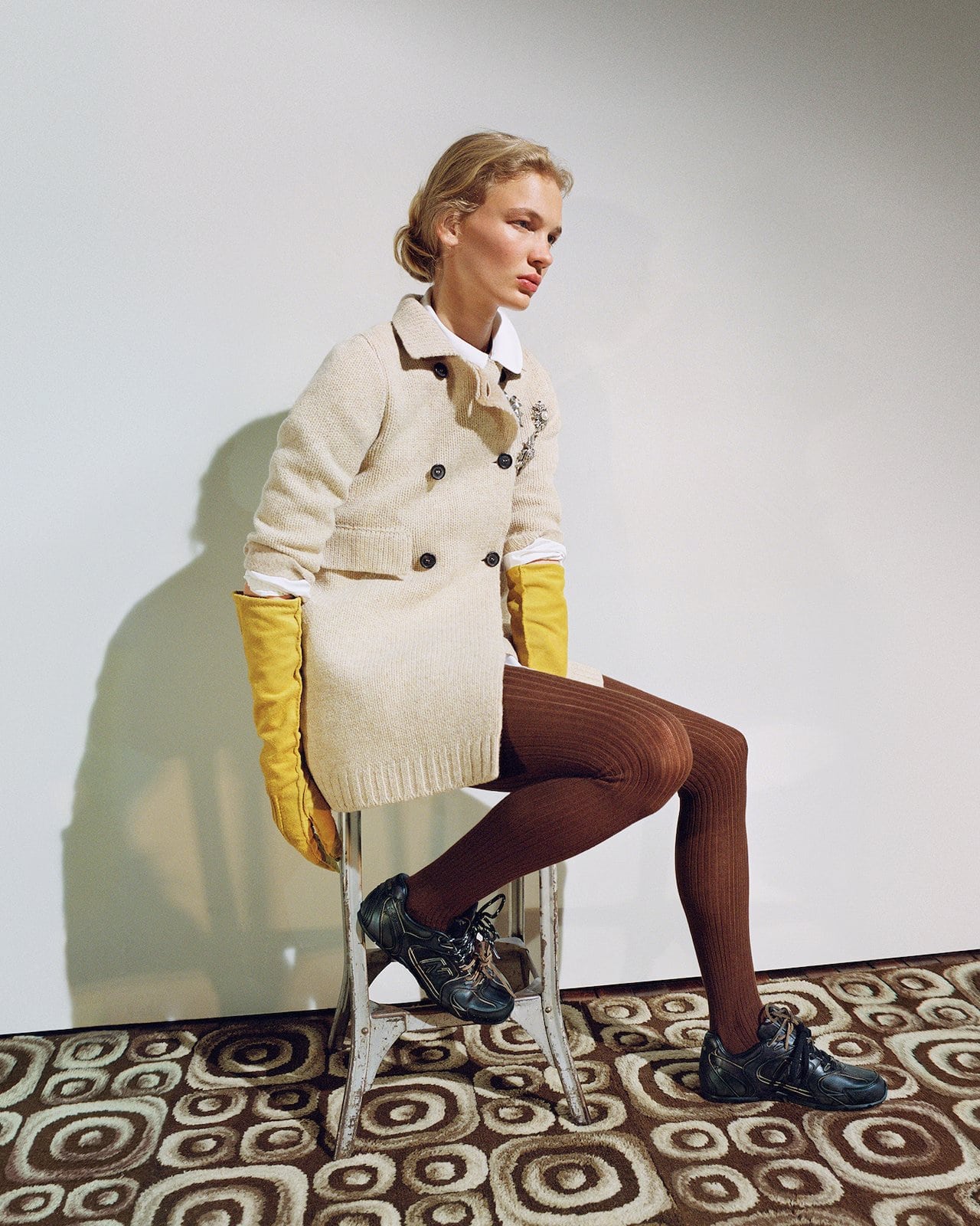 Model wearing cream double-breasted coat and yellow leather gloves sitting on a metal stool against a white wall.