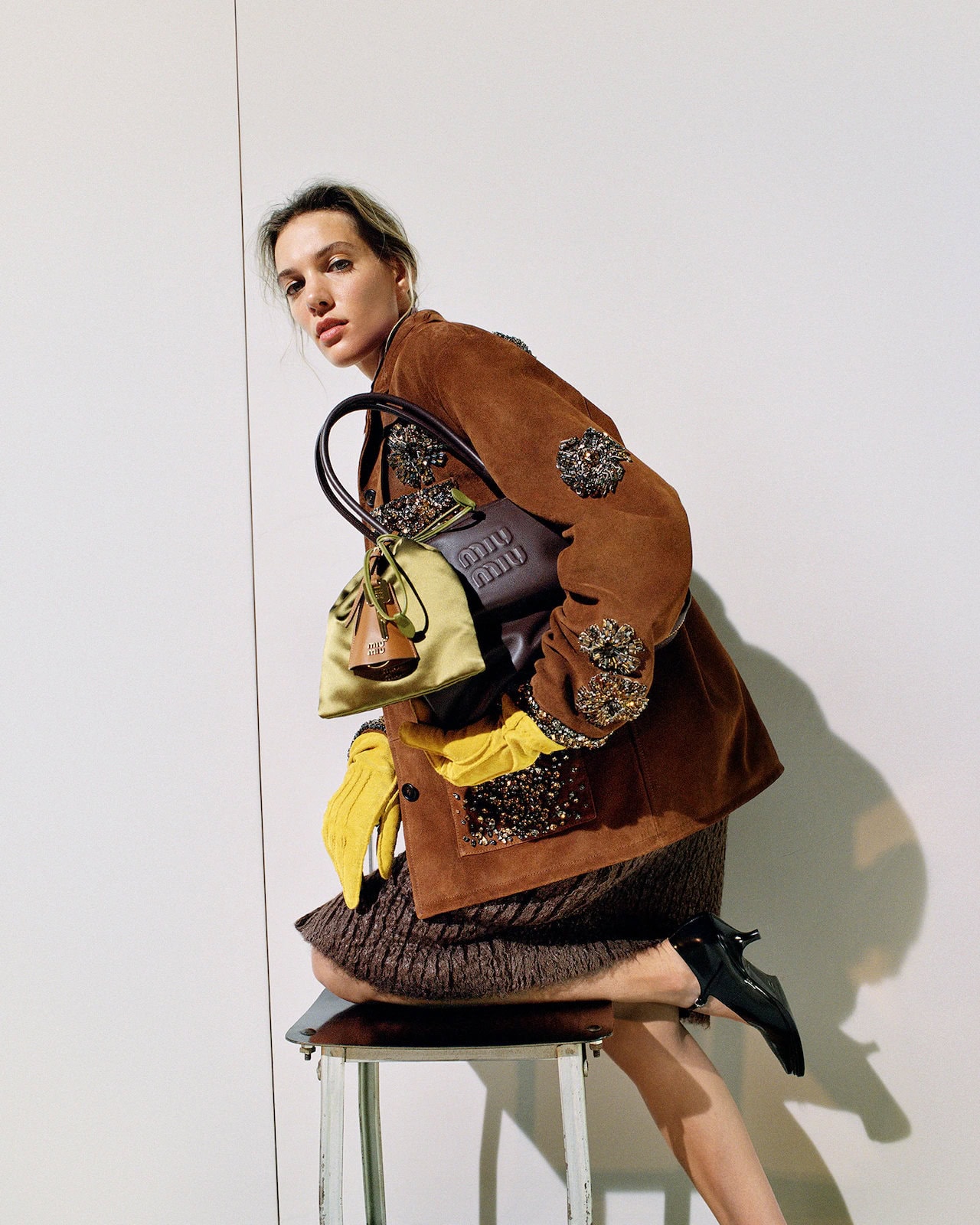 Fashion photography featuring an adult female model kneeling on a stool, holding multiple handbags, and wearing a brown jacket indoors.