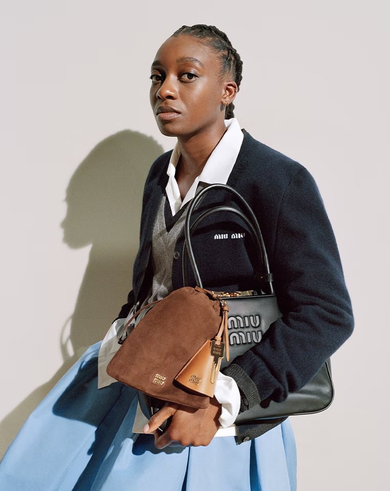 Model holding a brown suede pouch and black handbag wearing a dark blue cardigan over a white shirt and blue skirt.