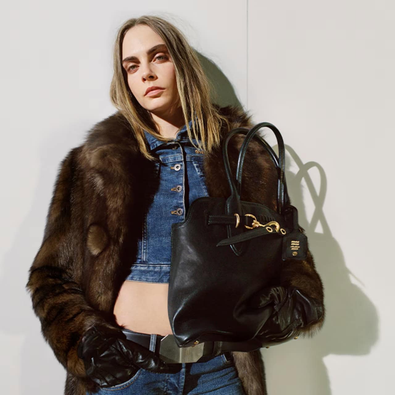 Fashionable woman posing in a fur coat, denim jacket, and blue jeans, holding a black leather handbag against a white backdrop.