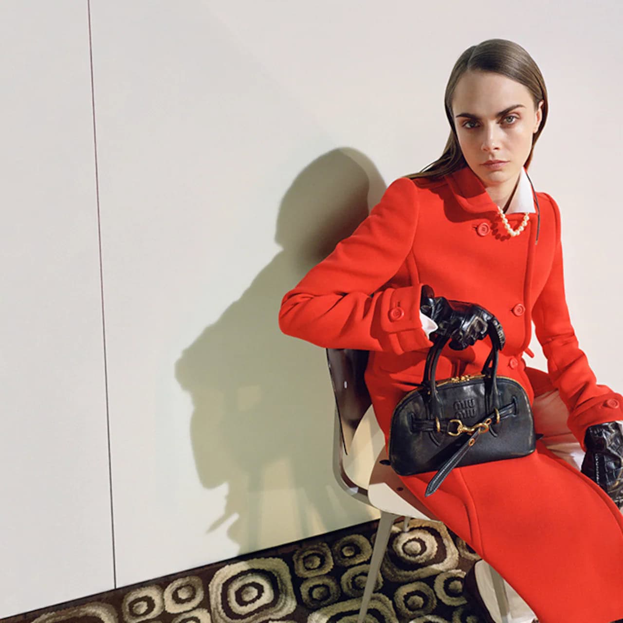 Model wearing a vibrant red coat and black leather gloves holding a dark handbag while seated on a chair indoors.