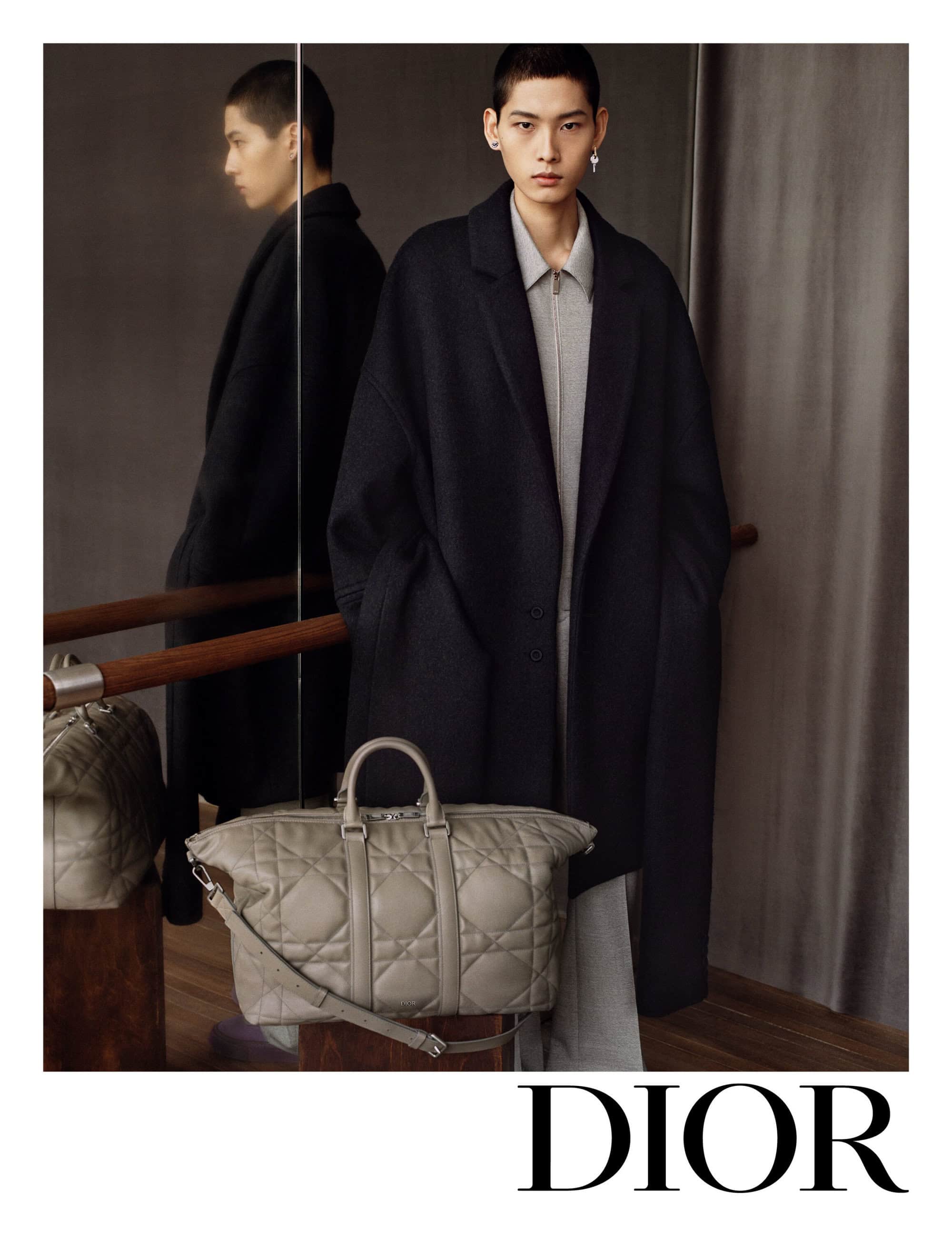 Fashion shot featuring a young adult male model in a coat, paired with a quilted grey leather duffel bag, presented in a photo studio.