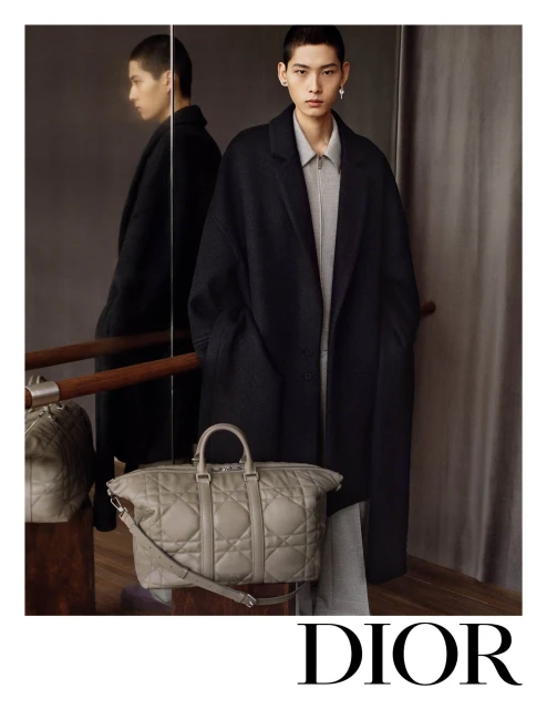 Fashion shot featuring a young adult male model in a coat, paired with a quilted grey leather duffel bag, presented in a photo studio.