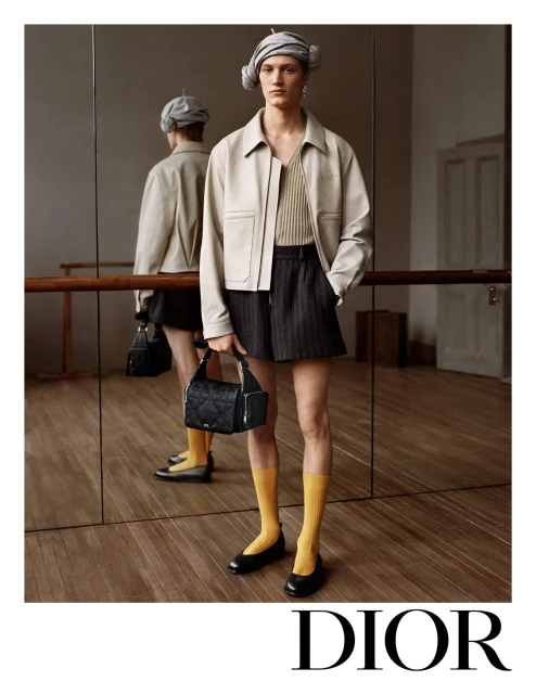 Fashion editorial shot of a slender adult wearing a jacket, shorts, socks and flats in a studio setting. Mirror reflection shows the model.