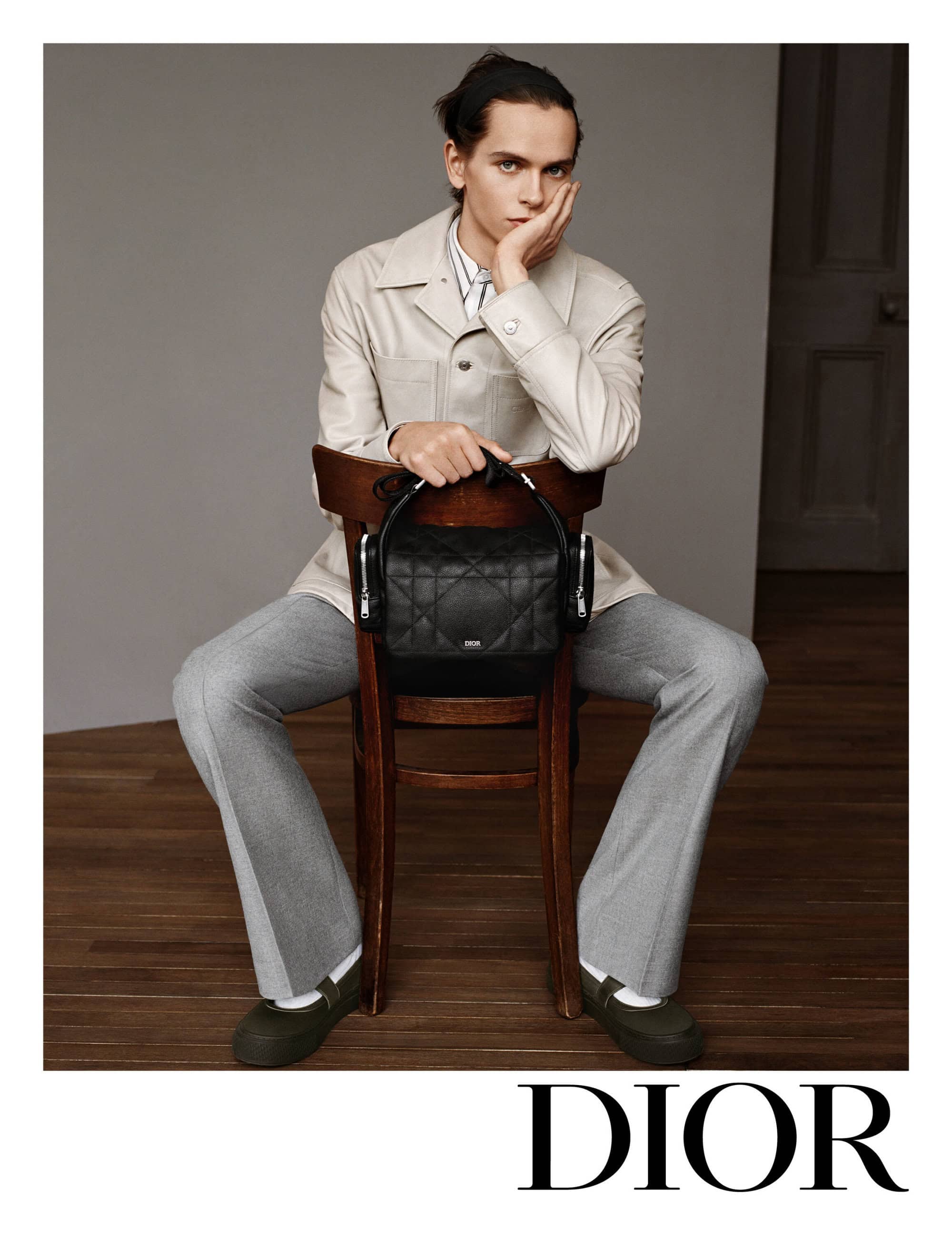 Model wearing a light beige jacket and grey trousers seated on a wooden chair holding a black quilted handbag against a neutral wall